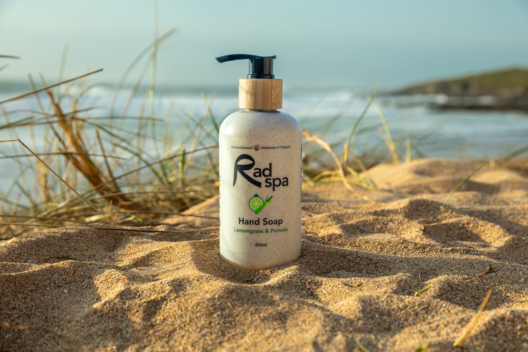 Hand soap bottle labeled 'Rad Spa' on a sandy beach with ocean in the background