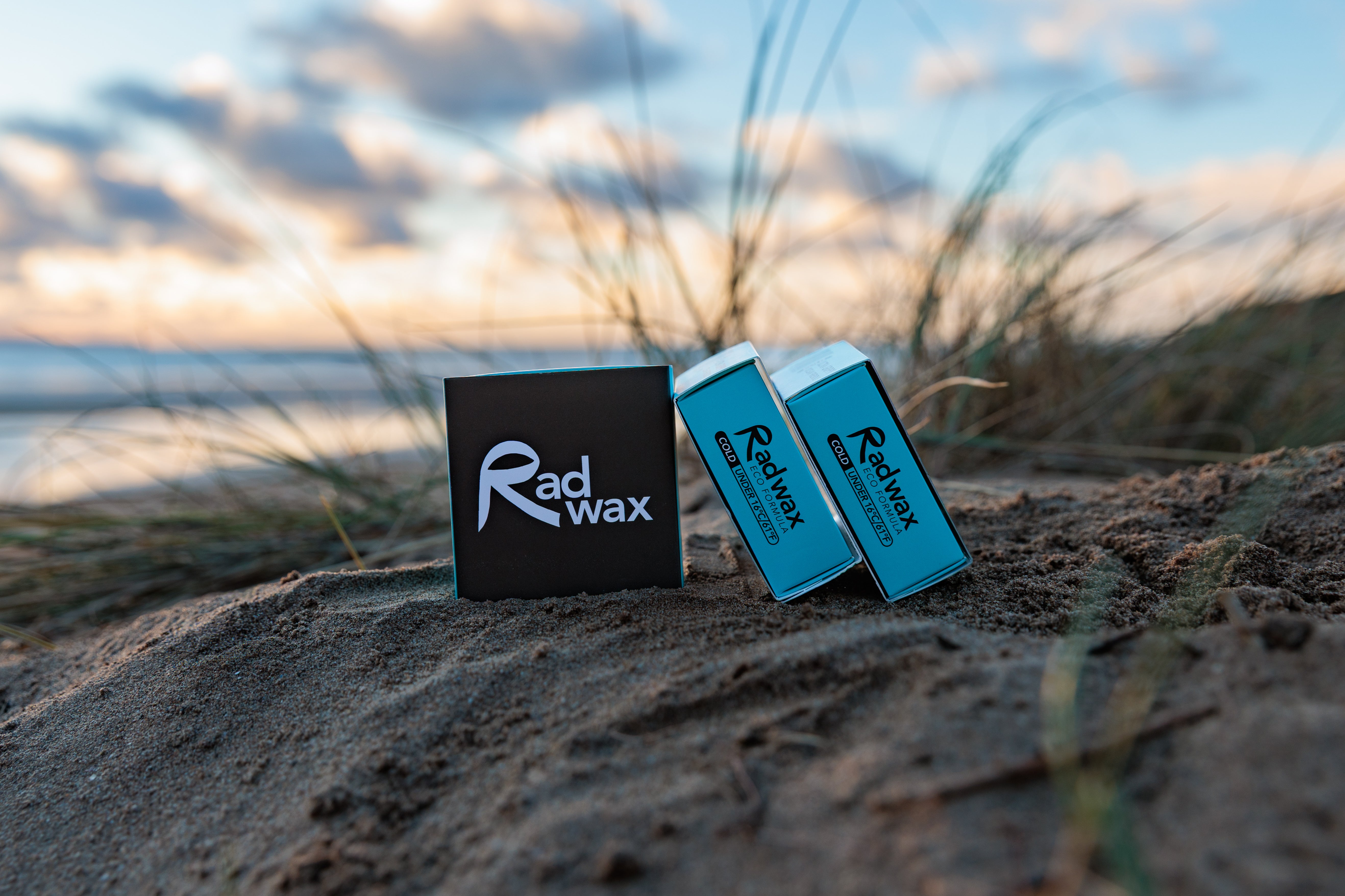 Rad Wax Cold Eco Surf Wax products on a sandy beach with grasses and water in the background