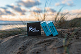 Rad Wax Cold Eco Surf Wax products on a sandy beach with grasses and water in the background