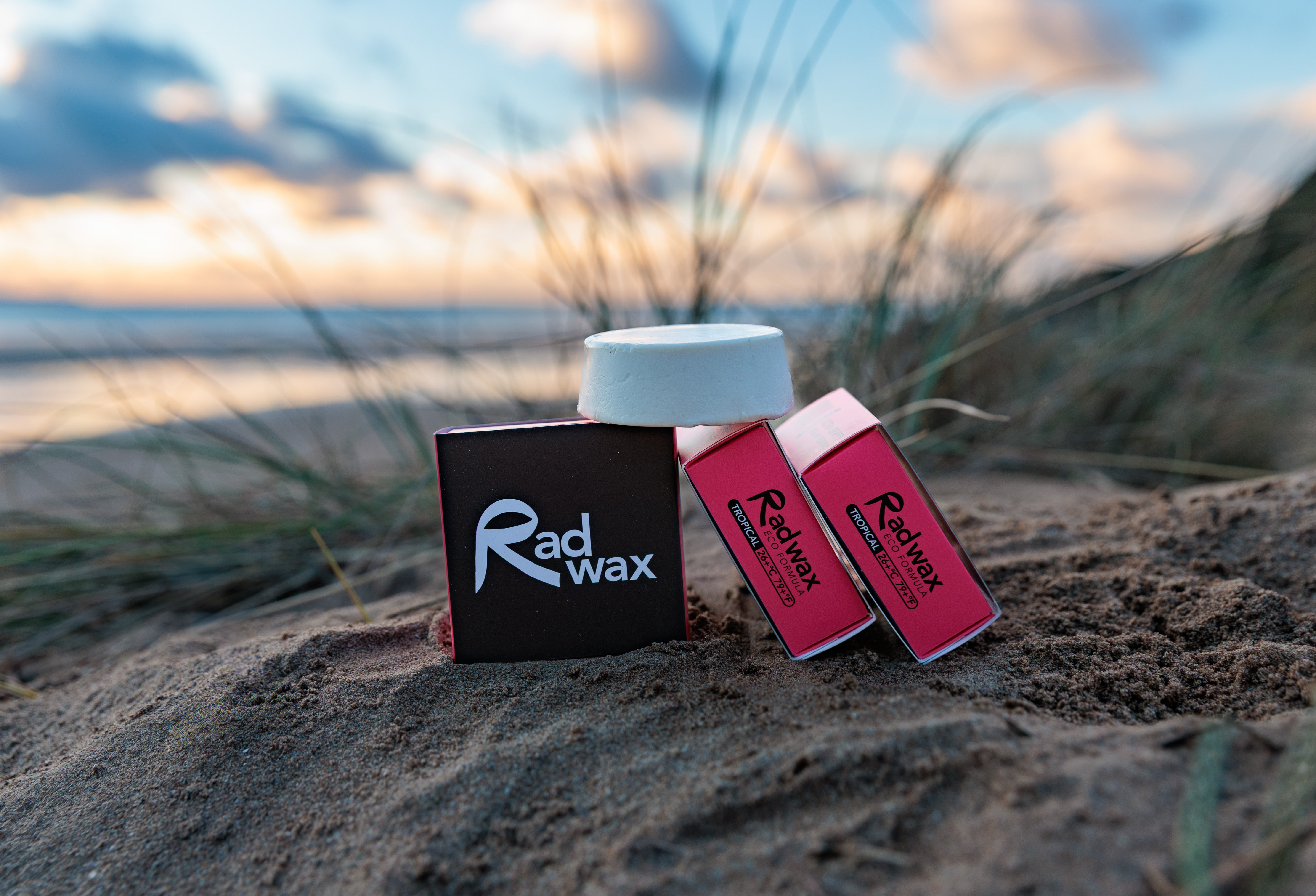 Rad Wax Tropical Eco Surf Wax on a sandy beach with grass and water in the background