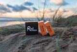 Rad Wax Warm Water Eco Surf Wax on a sandy beach with grass and water in the background